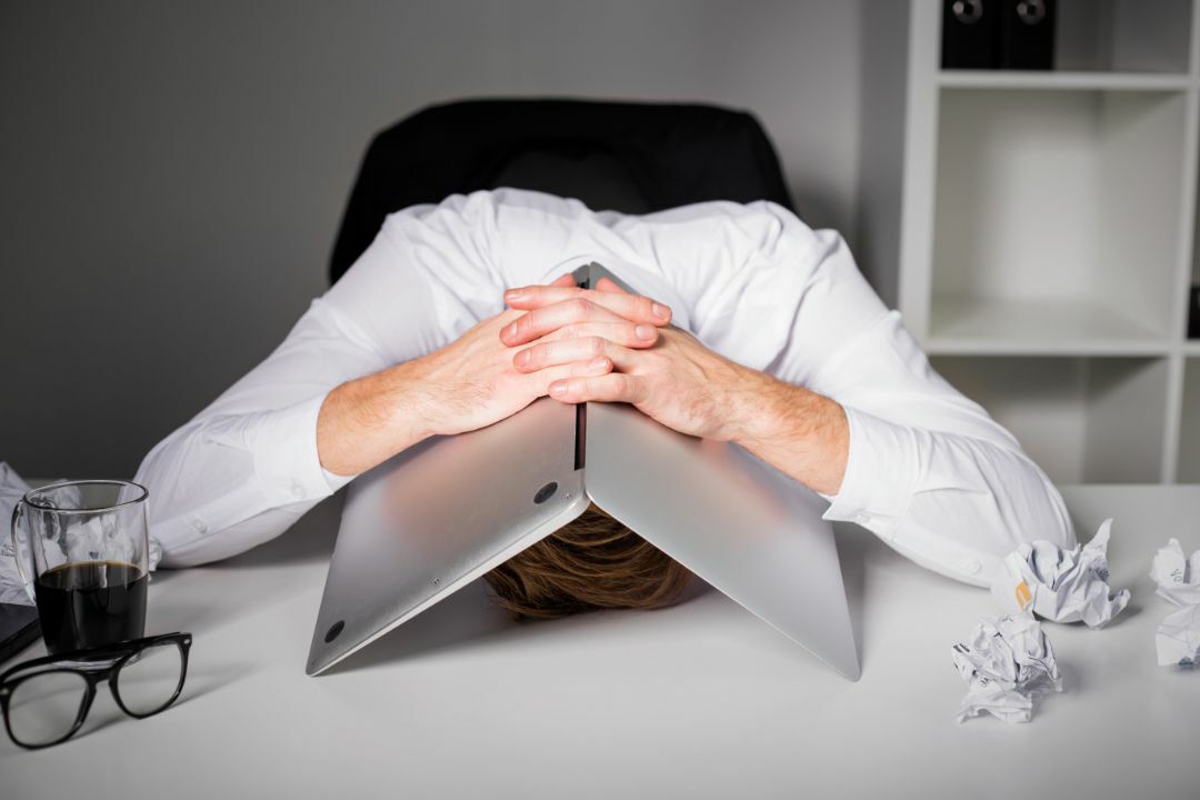 Verzweifelter Mann am Schreibtisch unter Laptop, da Fehler beim Immobilienverkauf gemacht wurden