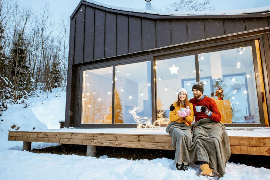 Mann und Frau sitzen im Winter mit Pudelmütze und Teetasse unter einer Wolldecke vor ihrem verschneiten Haus